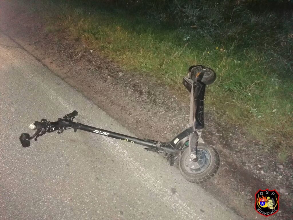 Uszkodzona hulajnoga elektryczna leży na poboczu drogi, obok roślinności.