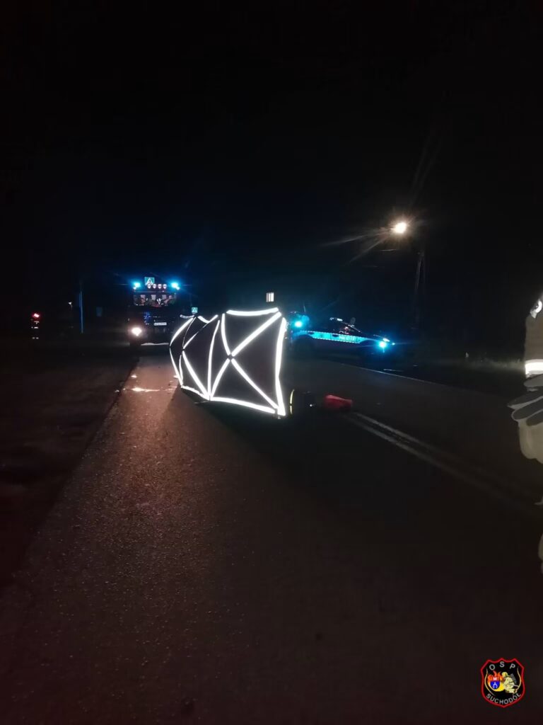 Na drodze leży odblaskowy trójkąt ostrzegawczy, obok widać pojazdy służb ratunkowych z włączonymi sygnałami świetlnymi.
