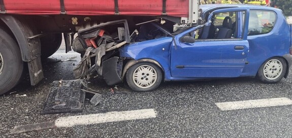 Niebieski samochód osobowy zgnieciony przez ciężarówkę na drodze, widać uszkodzoną część nadwozia.