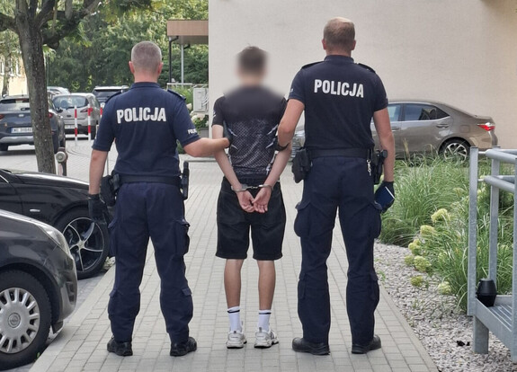 Dwóch policjantów w mundurach zatrzymuje mężczyznę z rękami związanymi z tyłu na chodniku w miejskim otoczeniu.