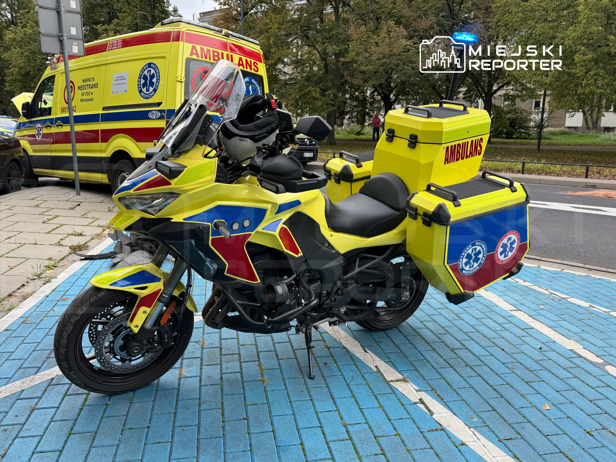 Motocykl służby ratunkowej w żółto-czerwonym malowaniu zaparkowany na niebieskiej kostce brukowej obok karetki.