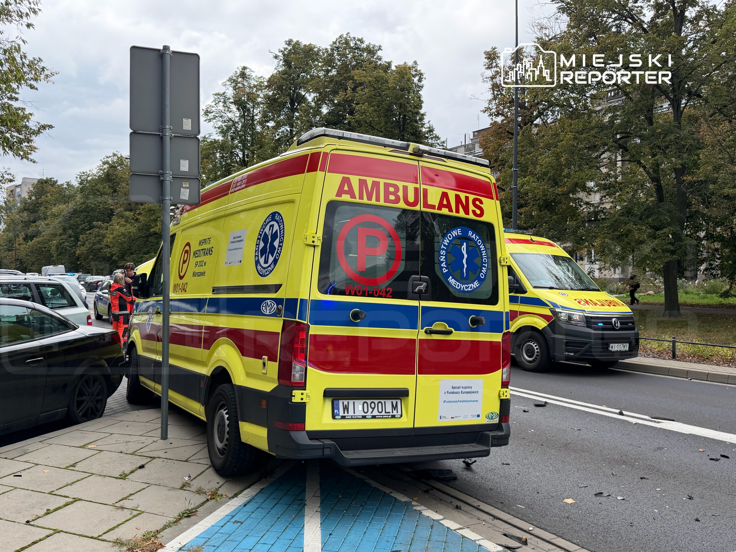 Ambulans z logo ratownictwa medycznego zaparkowany na chodniku, obok dwóch pracowników w strojach ochronnych.