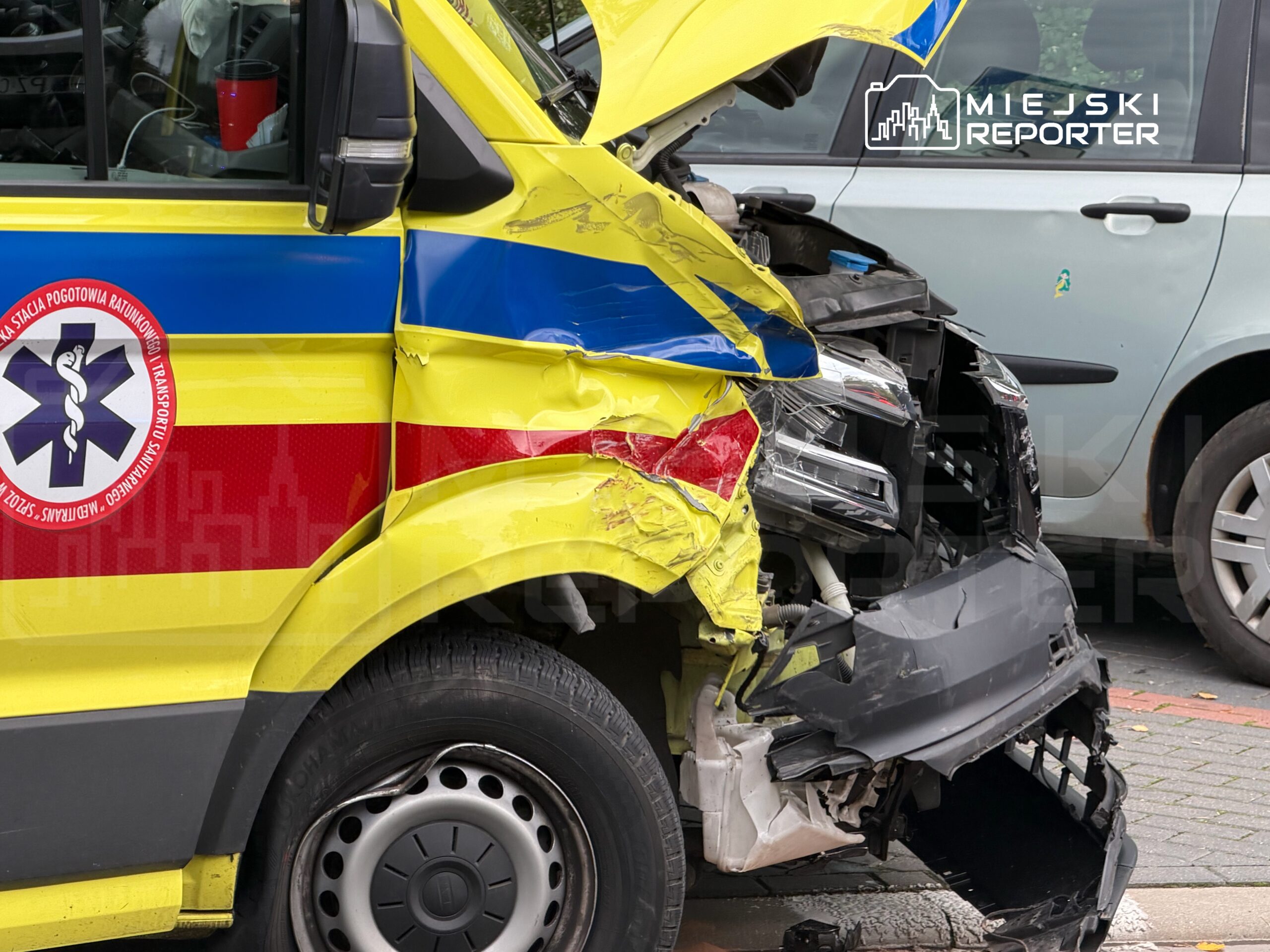 Ambulans z widocznymi uszkodzeniami bocznymi stoi na parkingu, obok innych zaparkowanych pojazdów.