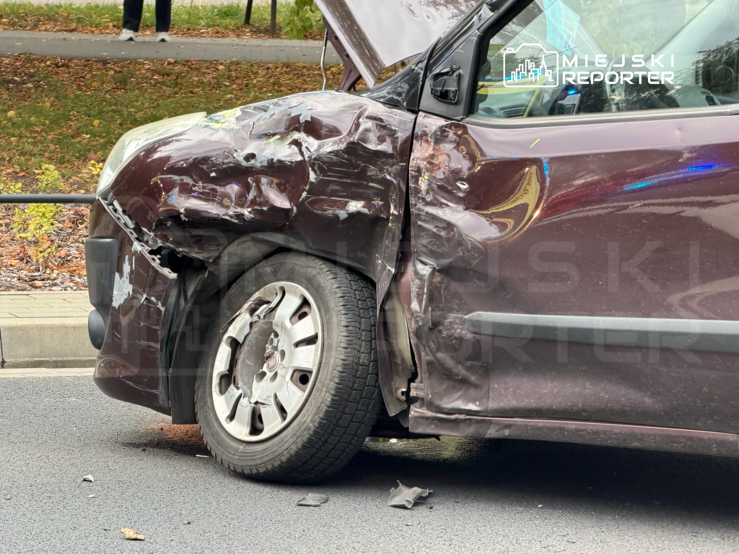 Uszkodzony samochód osobowy z wgniecioną przednią częścią stoi na jezdni, obok chodnika z zielenią.