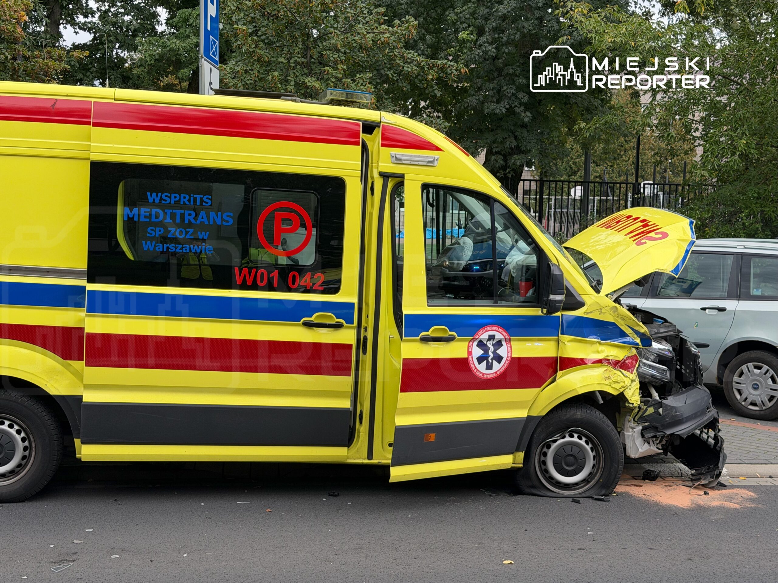 Ambulans z uszkodzoną przodu stoi na drodze, z otwartą maską, w otoczeniu parkowanych samochodów.