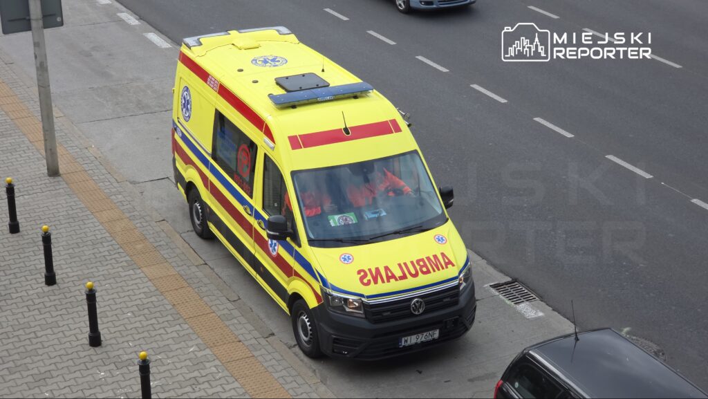 Żółta karetka pogotowia stoi na pasie ruchu, a w jej wnętrzu widoczni są ratownicy medyczni.
