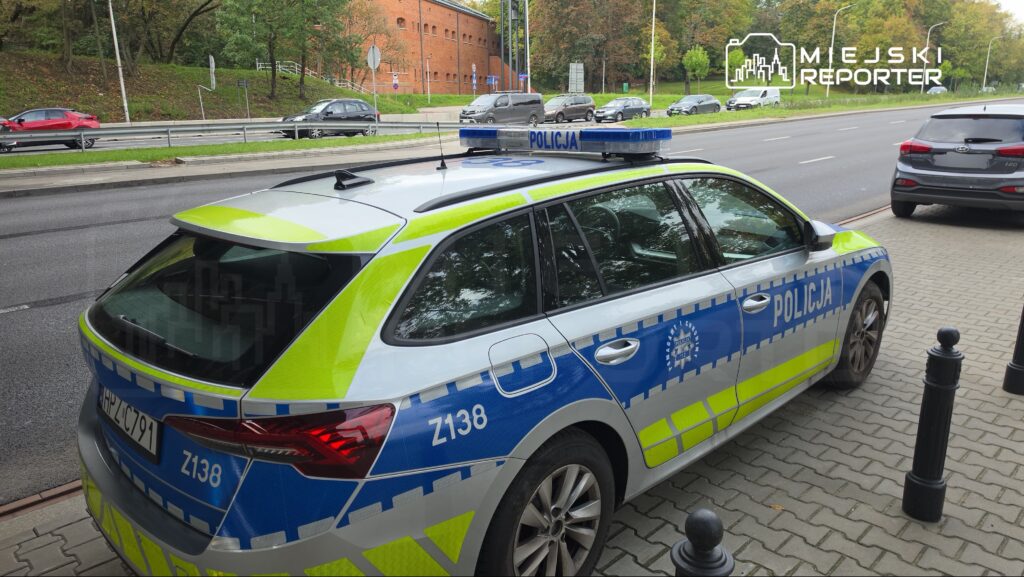 Policyjny radiowóz z niebiesko-zielonymi oznaczeniami zaparkowany na chodniku przy ruchliwej ulicy.