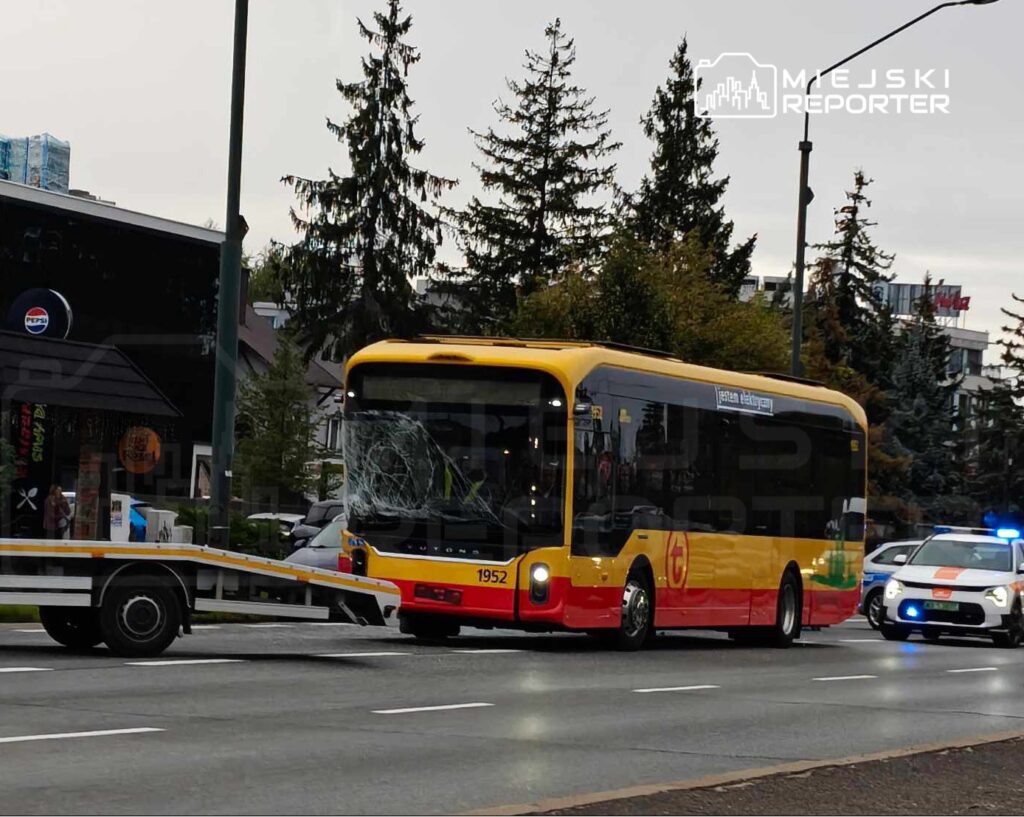 Zniszczony autobus komunikacji miejskiej holowany przez pojazd na ulicy, obok policyjny radiowóz.