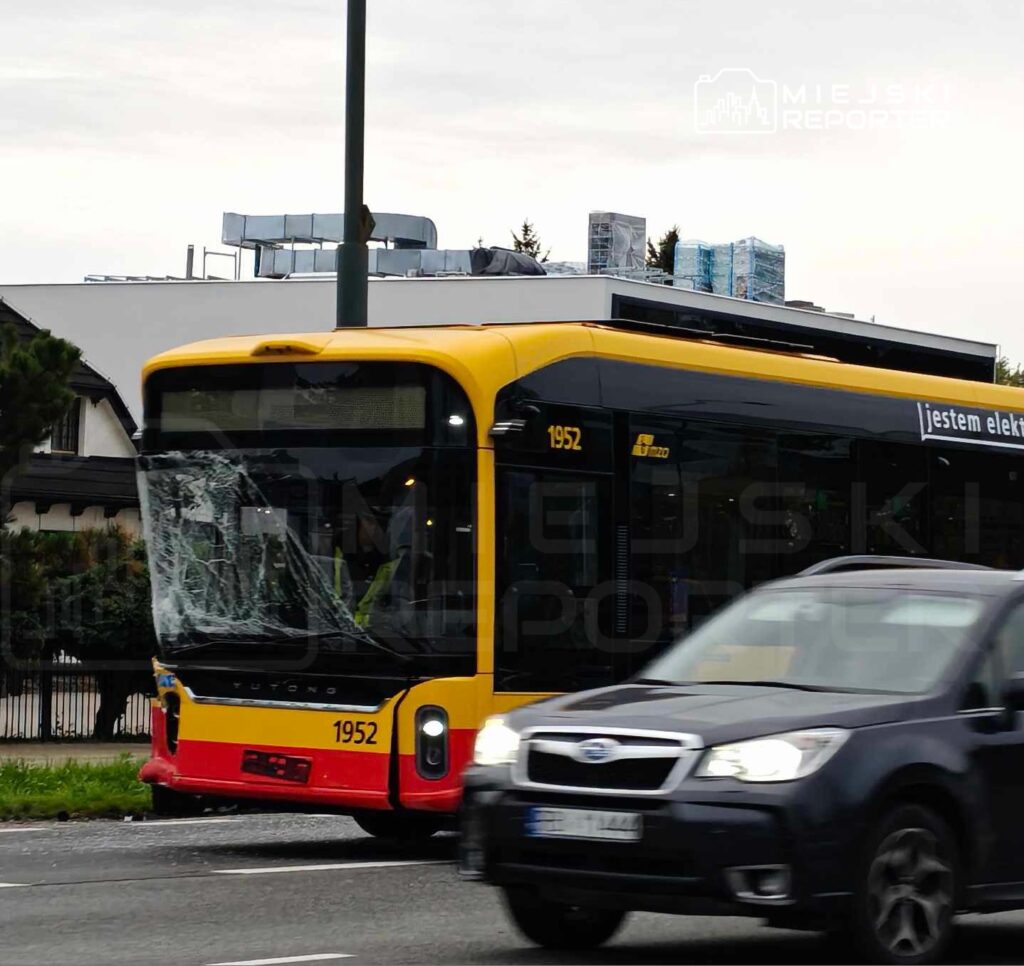Elektryczny autobus z widocznym uszkodzeniem szyby stoi na poboczu drogi, obok przejeżdżające samochody.