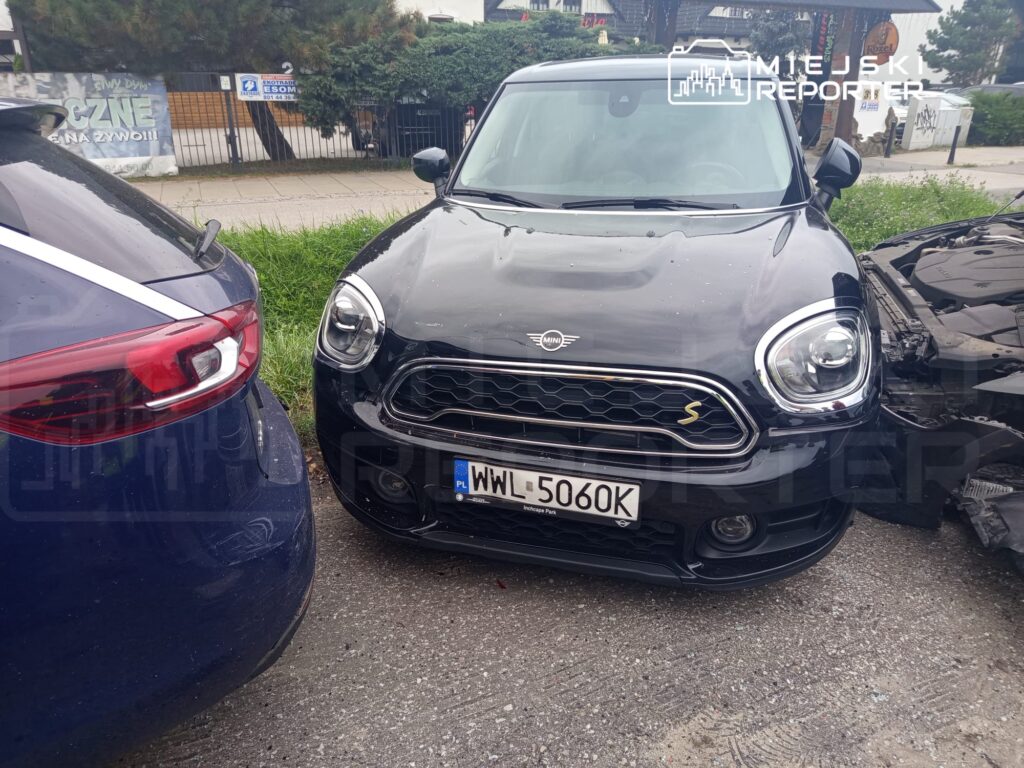 Czarny samochód osobowy marki Mini zaparkowany obok uszkodzonego pojazdu, w tle widoczny trawnik i ogrodzenie.