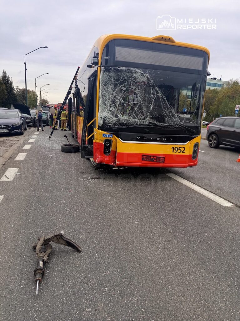 Uszkodzony autobus z rozbitym przednim szybą stoi na drodze, obok trwają prace ratunkowe. Na jezdni leży element pojazdu.