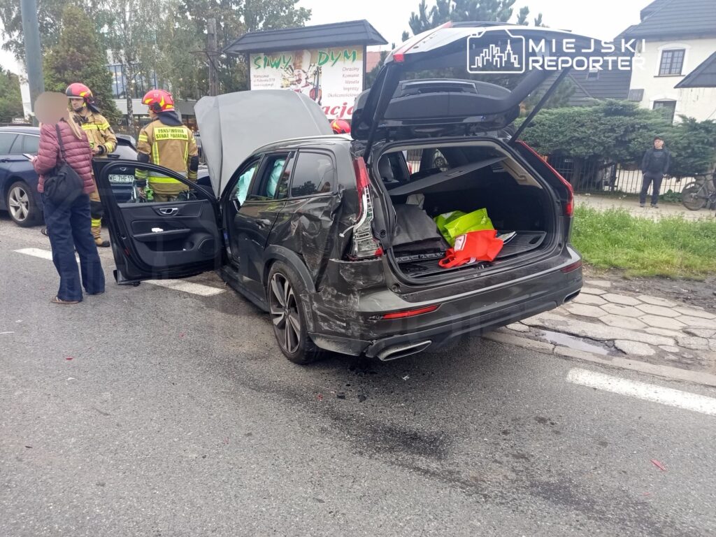 Strażacy udzielają pomocy przy rozbitej osobówce na jezdni, obok stoi zaniepokojona kobieta.