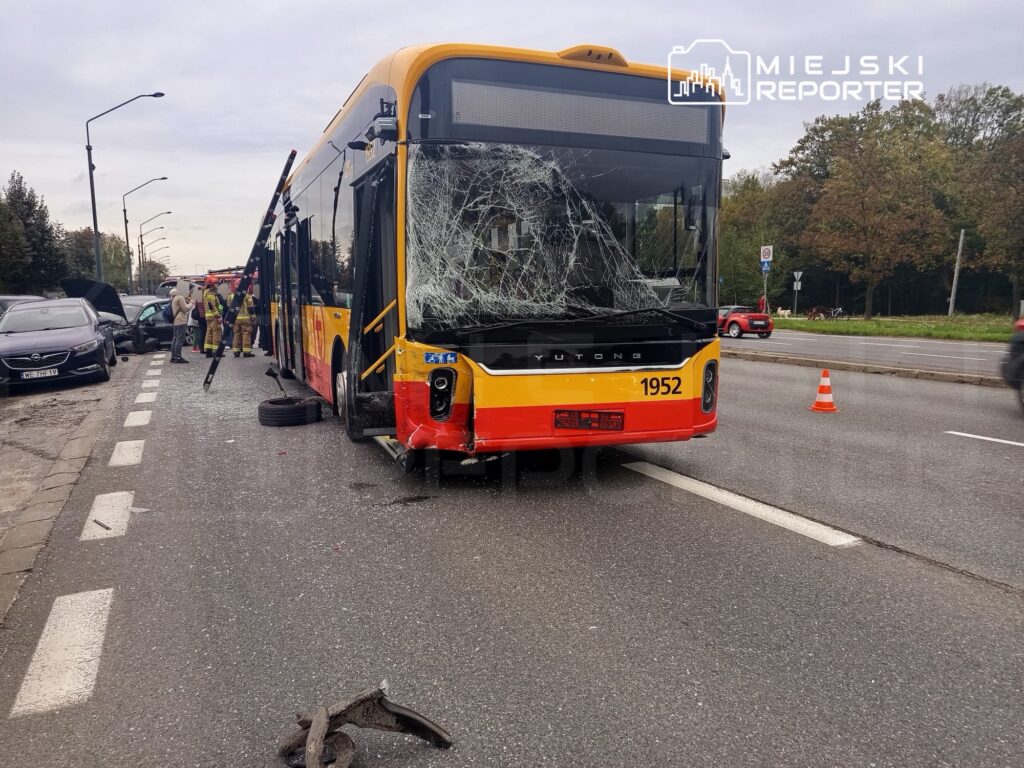 Zniszczony autobus z rozbitą szybą stoi na jezdni, obok pracują strażacy oraz ludzie przy uszkodzonych samochodach.