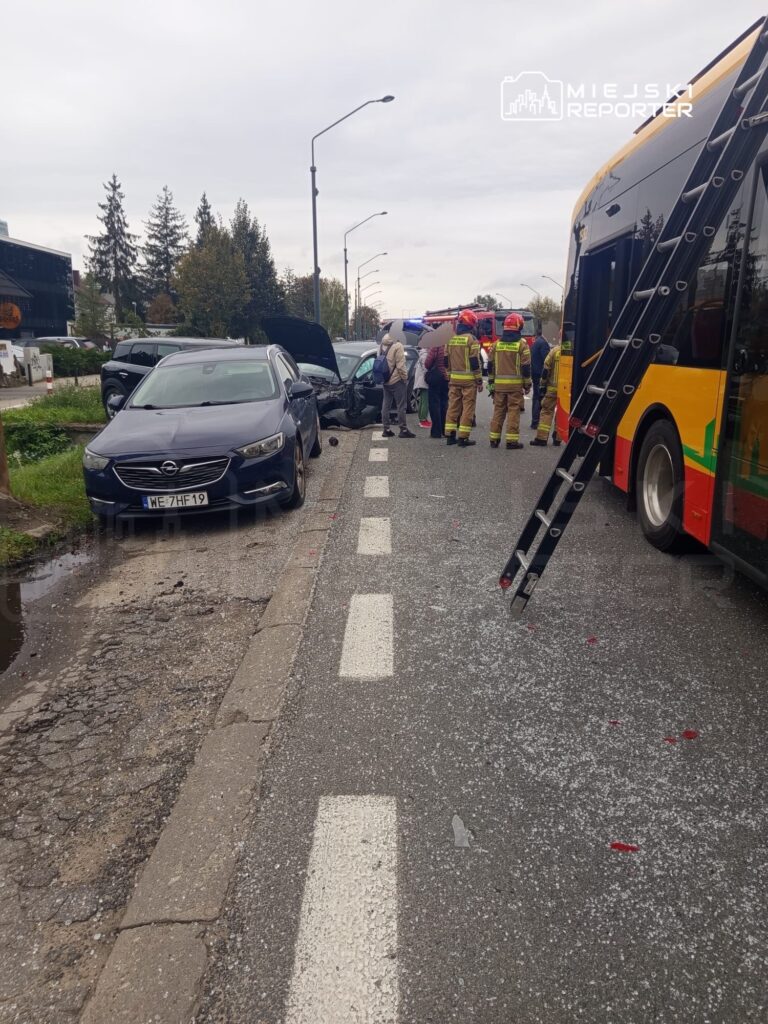 Na drodze znajduje się wypadek samochodowy, przy którym interweniują strażacy oraz kilka osób. W tle widoczny autobus.