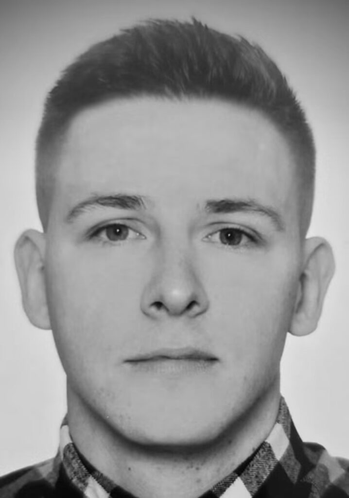 Mężczyzna z krótkimi włosami, w koszuli w kratę, patrzy prosto w obiektyw na białym tle.