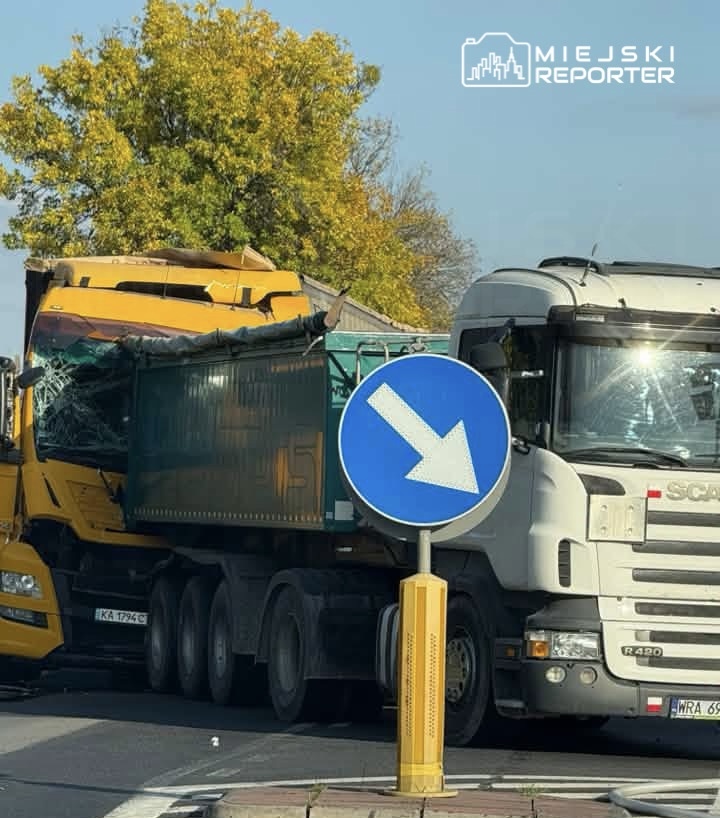 Dwa ciężarowe samochody, żółty i biały, stoją obok siebie za niebieskim znakiem nakazującym jazdę w prawo.