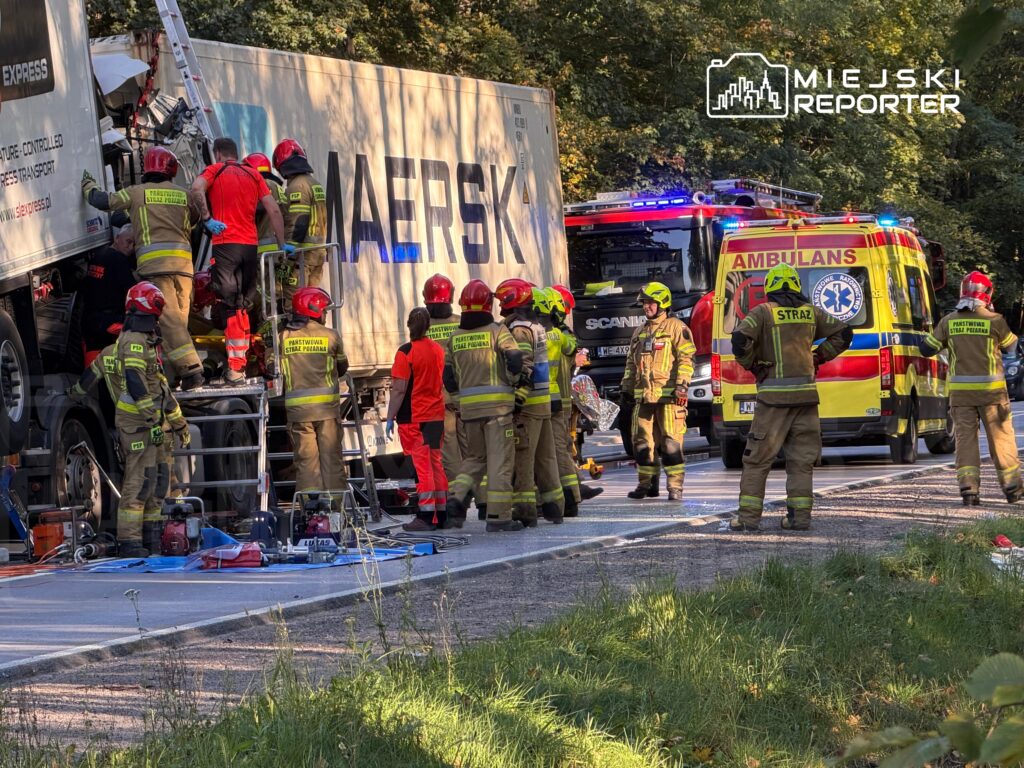 Strażacy i ratownicy pracują przy rozbitych ciężarówkach, obok karetki i wozu strażackiego na leśnej drodze.