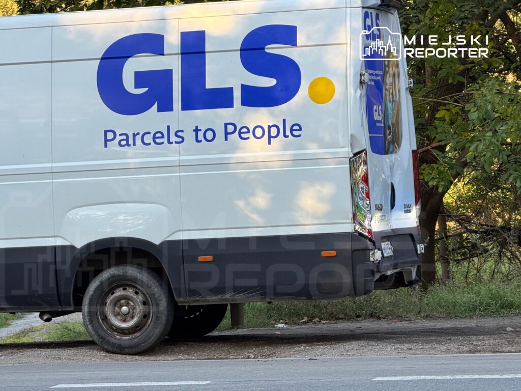 Biały samochód dostawczy GLS z napisem „Parcels to People” stoi przy drodze wśród drzew.