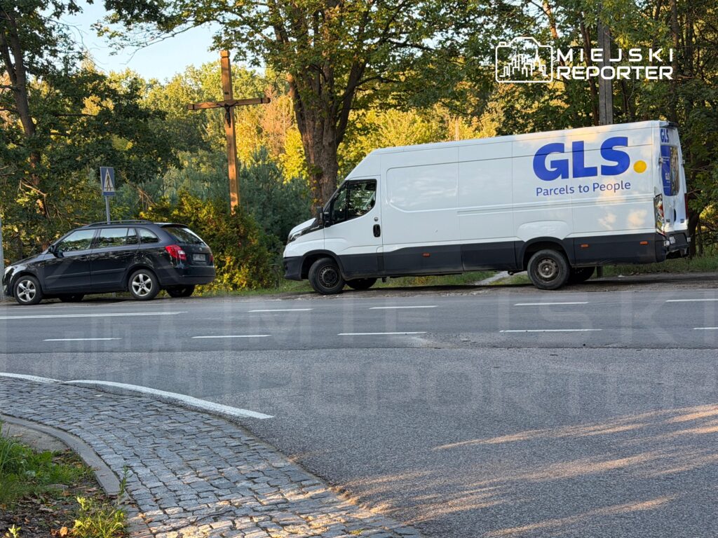 Biały furgon kurierski GLS i czarne kombi stoją przy drodze wśród drzew.