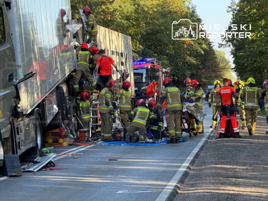 Strażacy i ratownicy pracują przy ciężarówce na drodze, obok stoją wozy strażackie