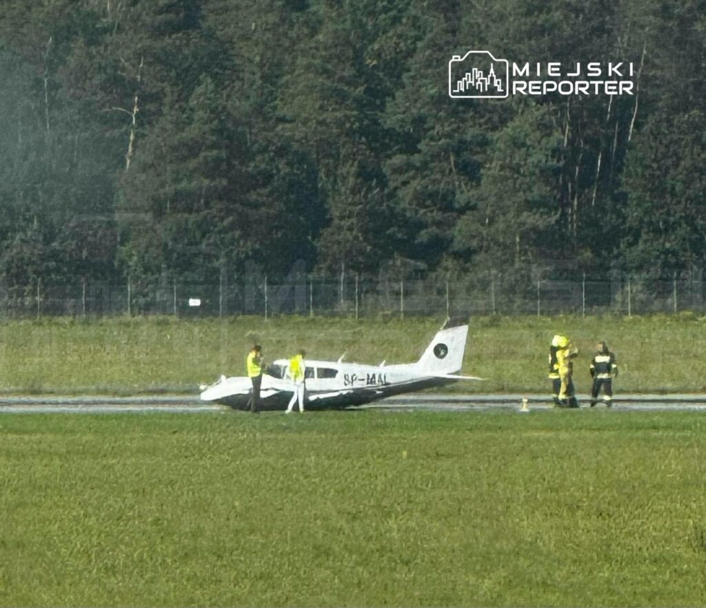 Mały samolot stoi na pasie lotniska, obok niego znajdują się osoby w kamizelkach odblaskowych i strażacy.