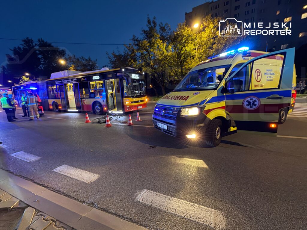 Autobus miejski i karetka pogotowia z włączonymi światłami stoją nocą na ulicy, wokół nich ustawiono pachołki.