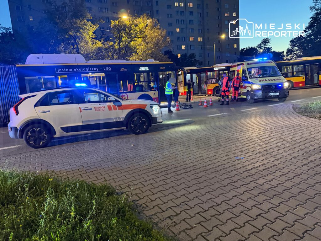 Karetka i samochód nadzoru ruchu stoją przy autobusach, a ratownicy pracują na ulicy po zmroku.