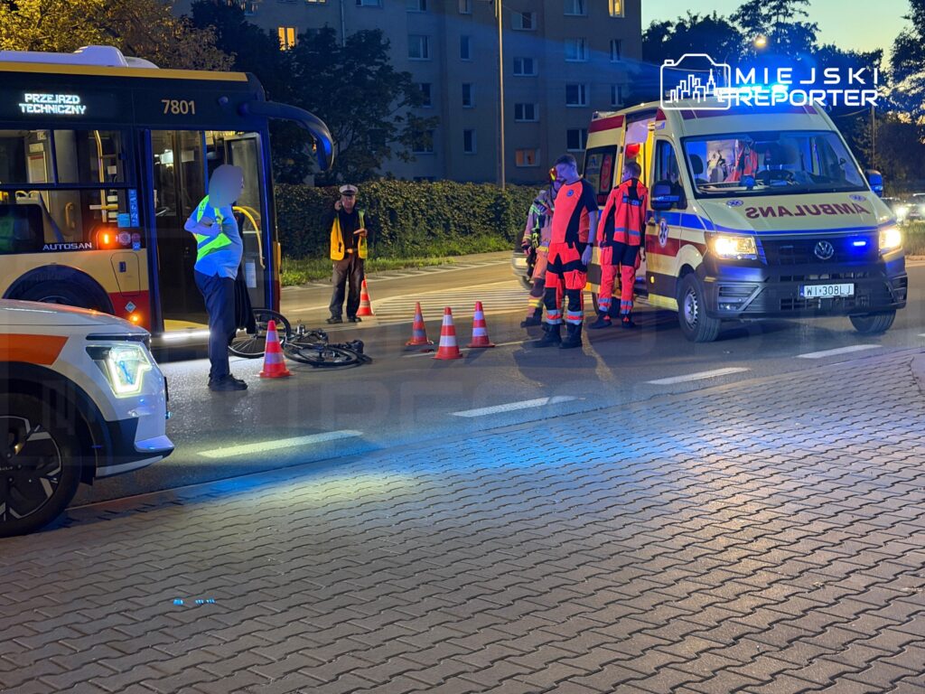 Ratownicy medyczni stoją przy karetce, autobusie i pachołkach na oświetlonej ulicy wieczorem.