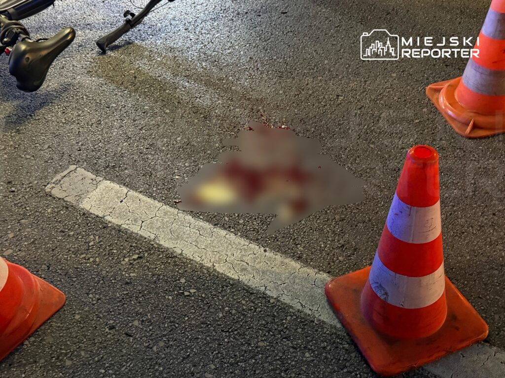 Pomarańczowe pachołki odgradzają rozmazane miejsce na jezdni obok roweru.