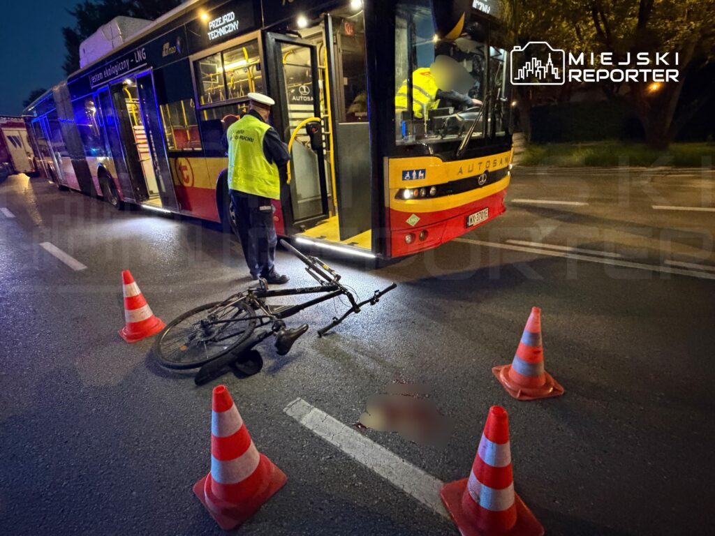 Przewrócony rower leży na jezdni obok autobusu, a policjant zabezpiecza miejsce zdarzenia pachołkami.
