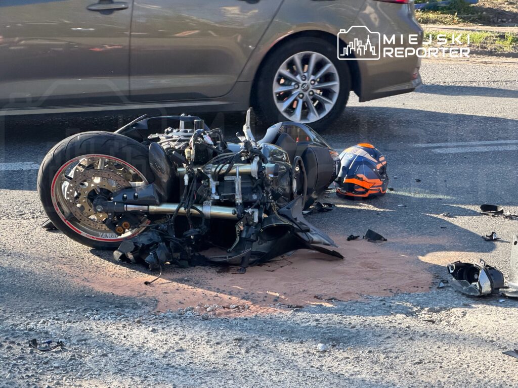Rozbity motocykl i kask leżą na jezdni obok zaparkowanego samochodu; wokół widać rozrzucone części i plamę płynu.
