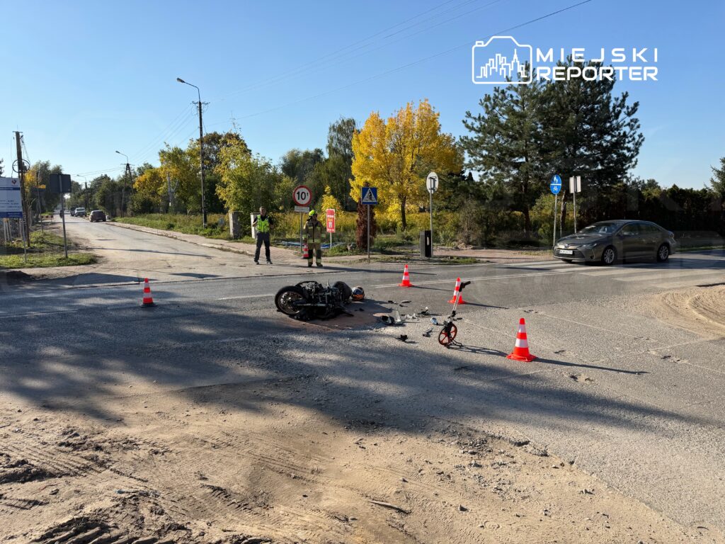 Przewrócony motocykl i rozsypane szczątki na skrzyżowaniu, zabezpieczone pachołkami; obok stoją służby ratunkowe.