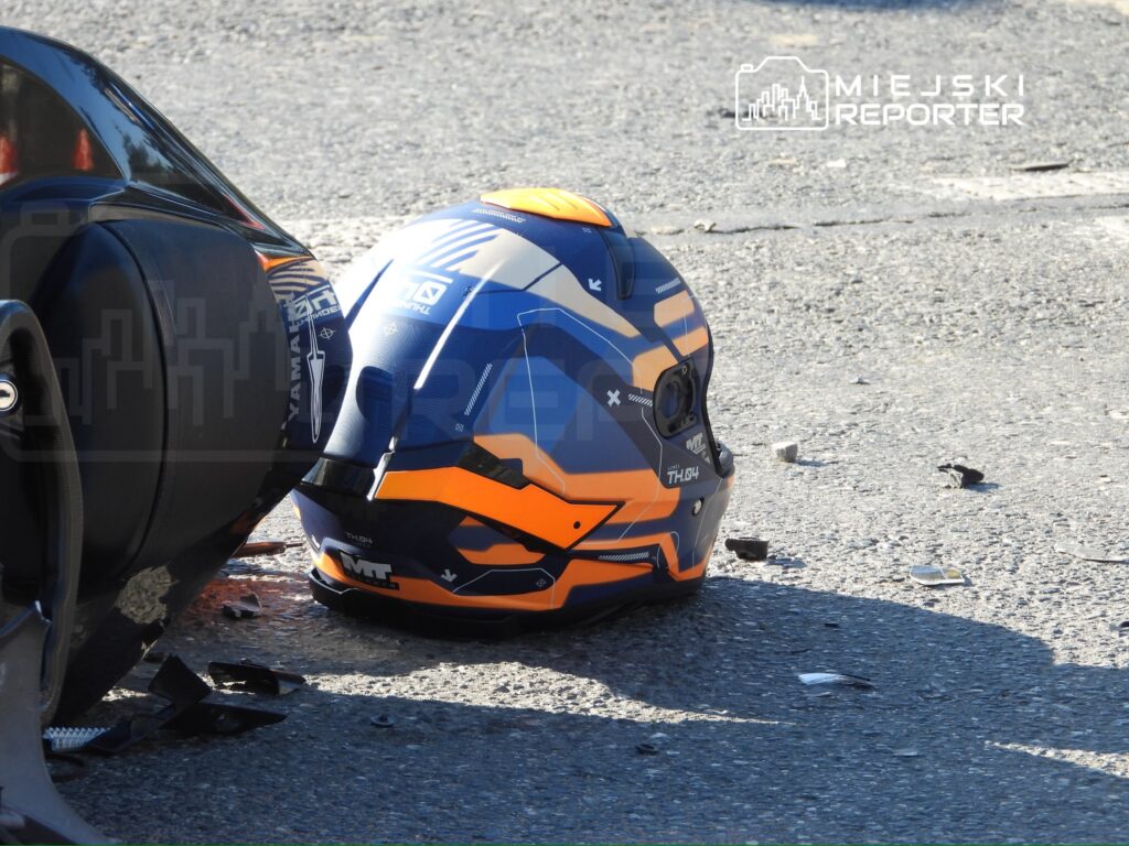 Niebiesko-pomarańczowy kask motocyklowy leży na jezdni obok uszkodzonego samochodu i odłamków.