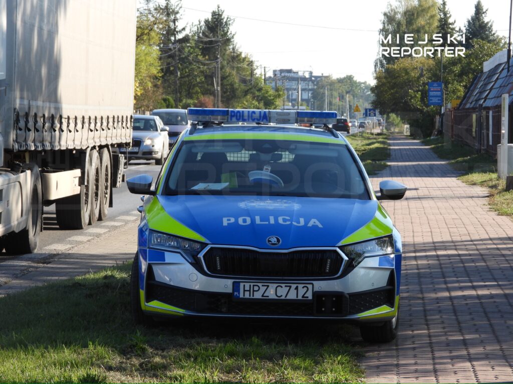 Oznakowany radiowóz policyjny stoi przy chodniku, obok ciężarówki i samochodów na miejskiej ulicy.