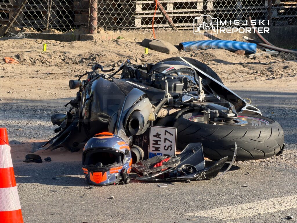 Uszkodzony motocykl leży na boku na jezdni, obok niego leży kask, a miejsce zabezpiecza pachołek drogowy.