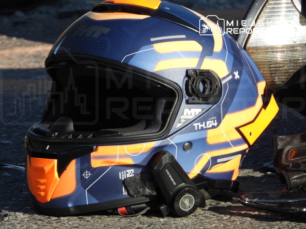 Niebiesko-pomarańczowy kask motocyklowy z zamontowaną kamerą leży na kamiennej nawierzchni obok motocykla.