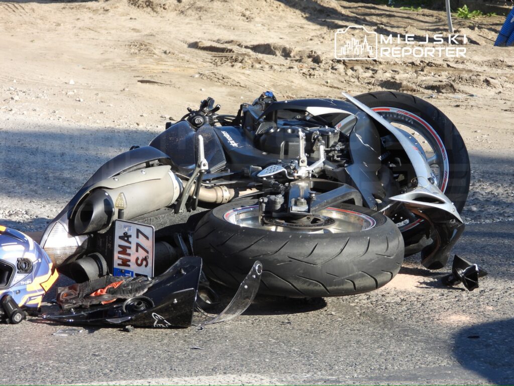 Przewrócony czarny motocykl leży na boku na asfaltowej drodze przy piaszczystym poboczu.