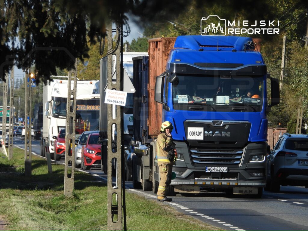 Strażak w ubraniu ochronnym stoi przed niebieską ciężarówką MAN z kontenerem na drodze, między samochodami.