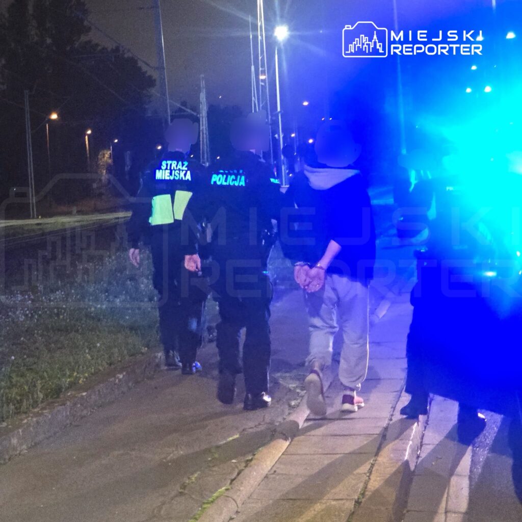 Funkcjonariusze straży miejskiej i policji prowadzą osobę w kajdankach chodnikiem nocą, przy błyskach niebieskich świateł