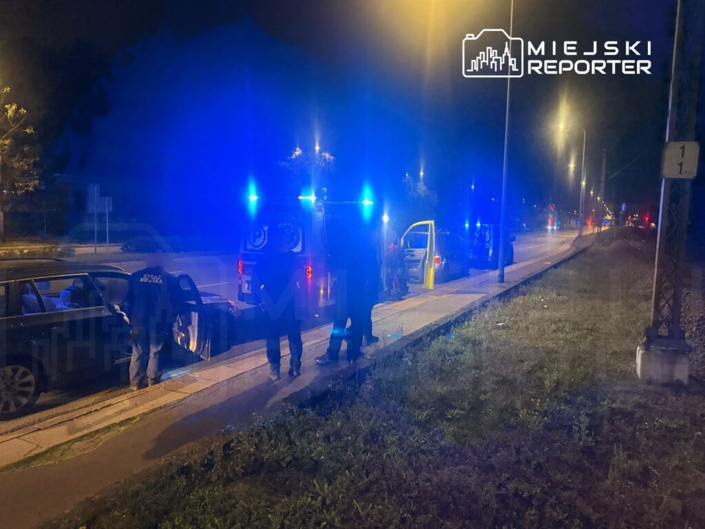 Policjanci stoją przy radiowozach z włączonymi niebieskimi światłami na nocnej ulicy.
