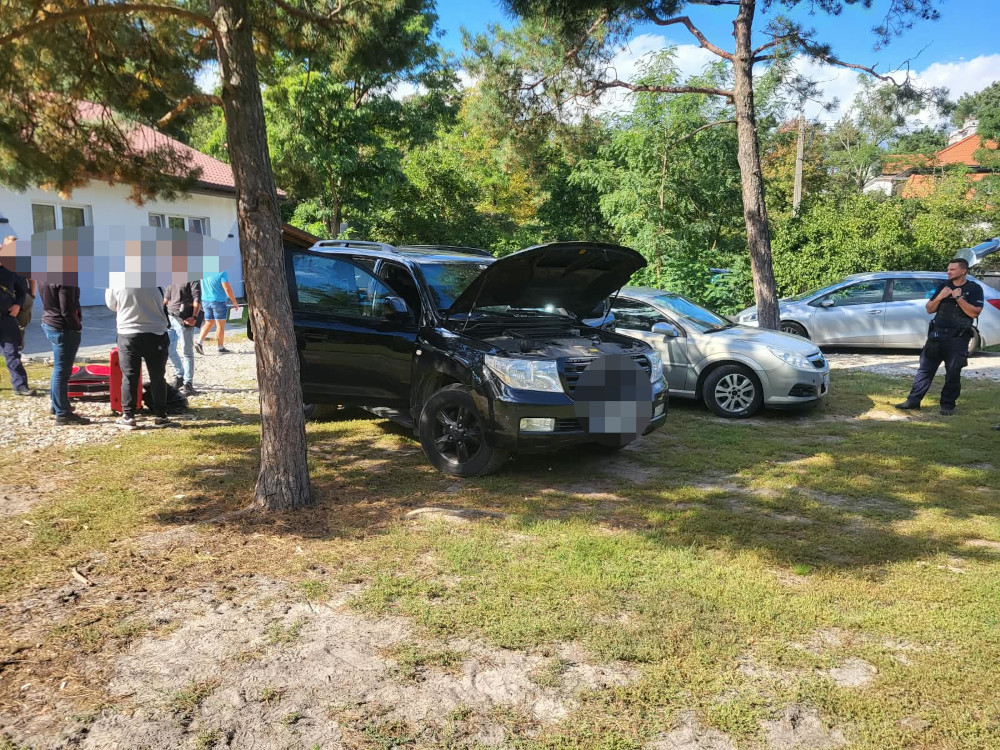 Czarny SUV z otwartą maską stoi na zaleszonym parkingu, obok zaparkowanych samochodów i grupy osób.