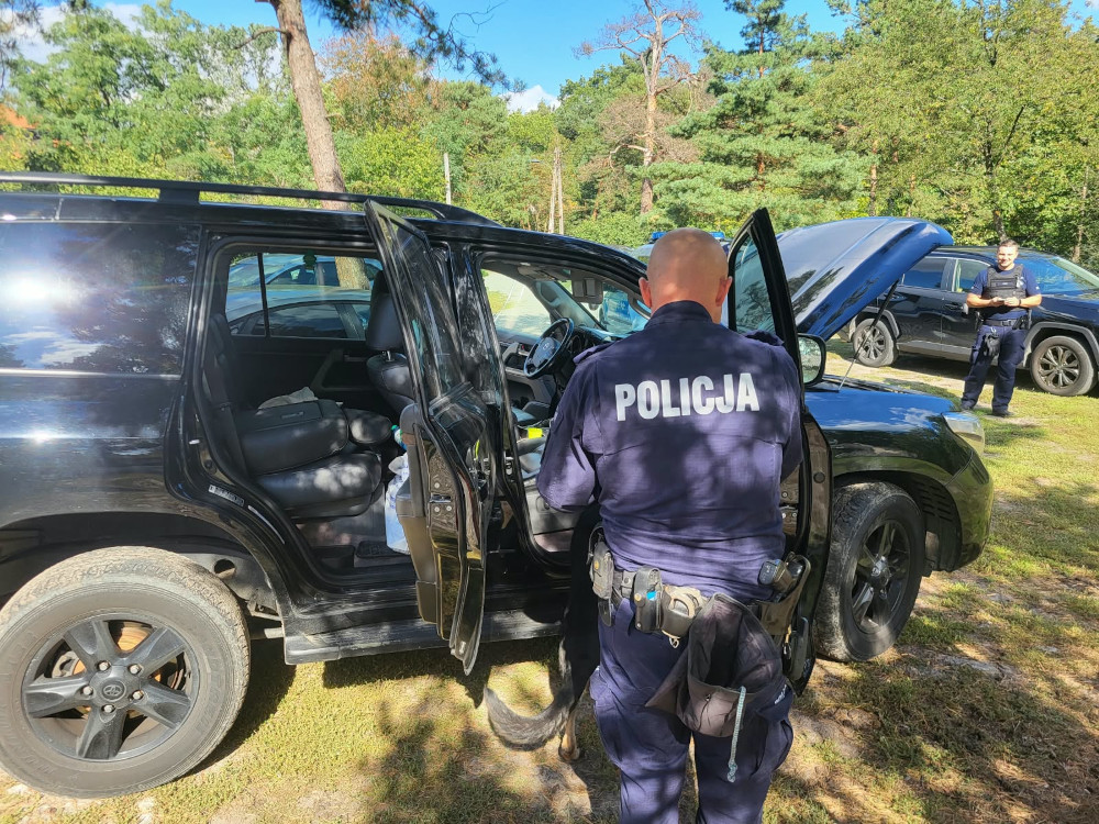 Policjant sprawdza wnętrze czarnego SUV-a z otwartymi drzwiami i maską na leśnym parkingu.