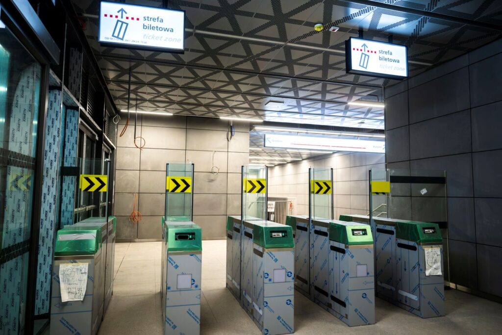 Nowoczesne bramki biletowe w strefie biletowej nowego tunelu linii metra M2, z oznaczeniami kierunkowymi.