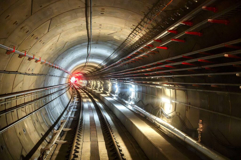 Tunel metra M2 z widocznymi torami i oświetleniem, prowadzący w kierunku czerwonego światła na końcu.