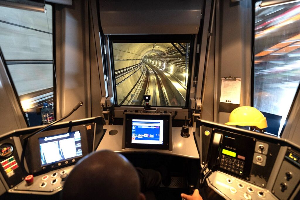 Maszynista w kabinie pociągu M2, prowadzący pojazd przez tunel z widokiem na zakręt torów.