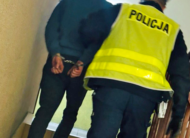 Mężczyzna w kajdankach prowadzony przez policjanta w kamizelce odblaskowej na korytarzu budynku.