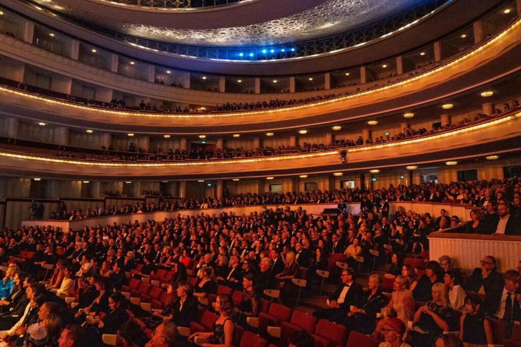 Publiczność zajmuje wielopoziomową widownię oświetloną ciepłym światłem w ozdobnym teatrze.