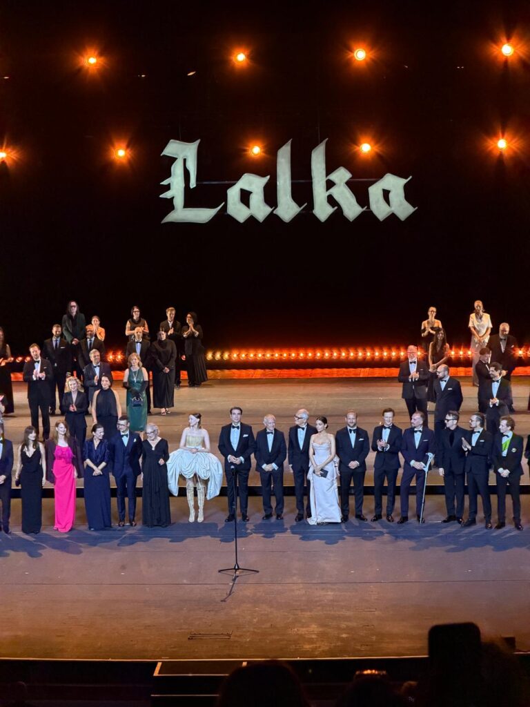 Aktorzy w eleganckich strojach kłaniają się na scenie pod napisem „Lalka” podczas uroczystej premiery spektaklu.