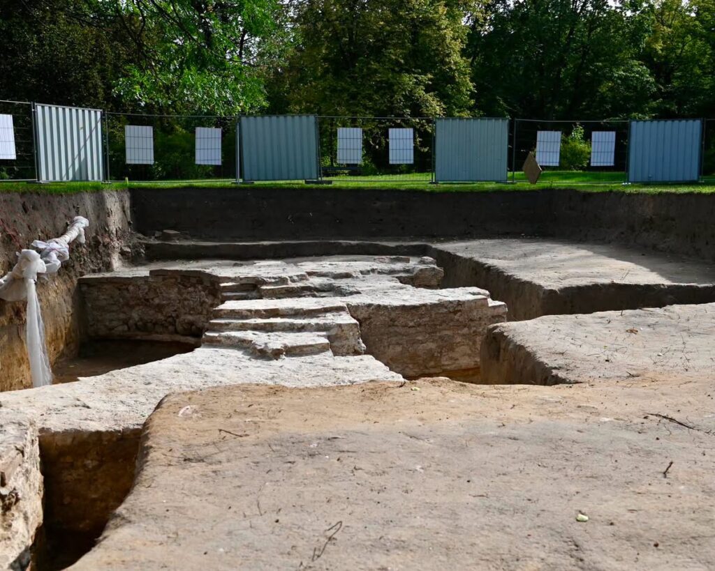 Wykopaliska archeologiczne z widocznymi schodami i ułożonymi kamieniami w otoczeniu ogrodzenia budowlanego.