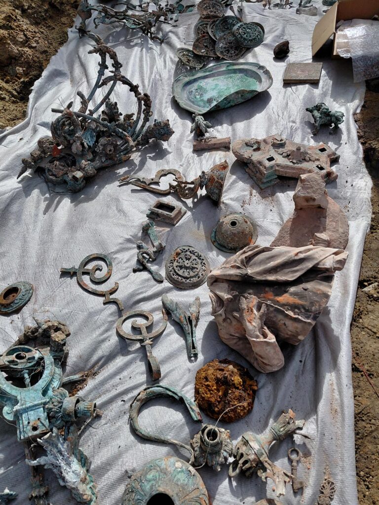 Zabytkowe przedmioty metalowe i ceramiczne leżą na białym płótnie, wśród nich fragmenty rzeźb i ozdobne elementy.